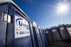 two rows of blue portable toilets with "United Site Services" logo lined up outdoors in a daytime setting two rows of blue portable toilets with "United Site Services" logo lined up outdoors in a daytime setting