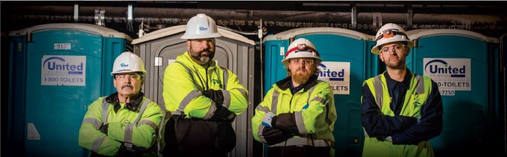 four men wearing a hard hats and hi-vis vests standing in front of a portable toilet outside