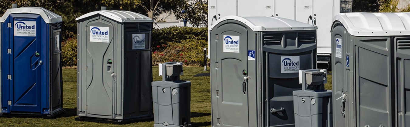 Row of portable toilets and hand washing stations on grass arranged against a background of bushes with a restroom trailer in the back