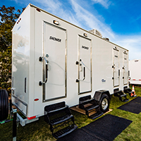 A portable shower trailer with labeled doors under a clear sky, parked on grass with small entry stairs and a black mat at each door