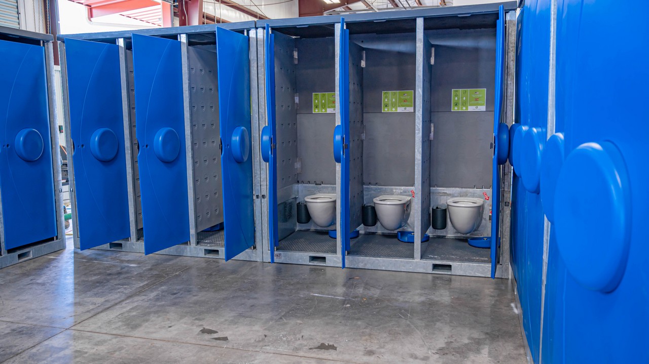 A row of open blue Hydroflow bathroom stalls with toilets inside an industrial building, showcasing privacy doors partly open.
