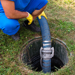 A worker in a blue uniform and yellow gloves operating a large hose to pump out a septic tank