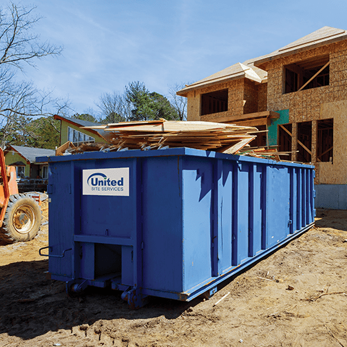 A blue roll-off dumpster filled with construction debris, in front of partially built wooden structures, under a clear sky