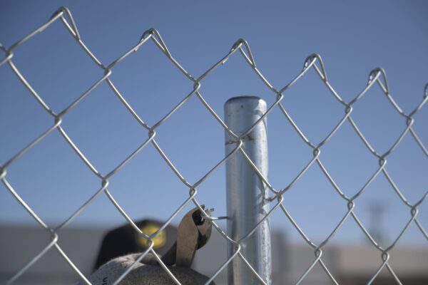 A chain-link fence with a focus on its interwoven metal wires, showing a blurred background featuring a metal pole
