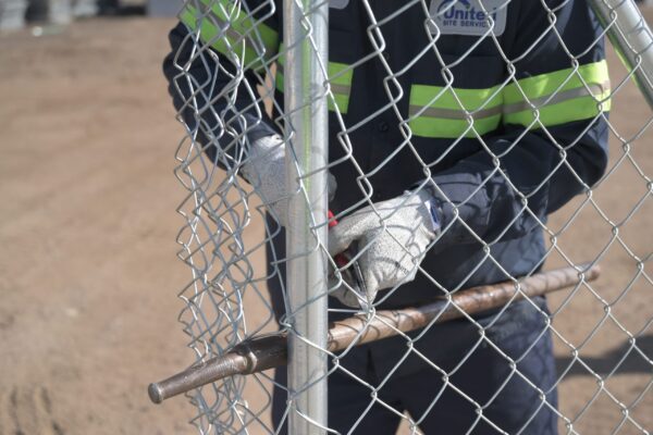 A worker in a reflective vest and gloves unlocking a chain-link fence gate, holding a metal bolt cutter