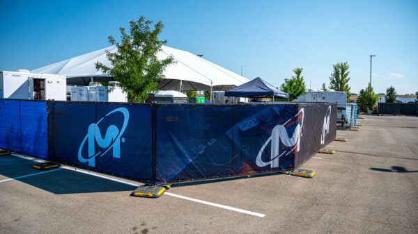 A large blue temporary fence with white graffiti-style artwork enclosing an area with a white tent visible in the background