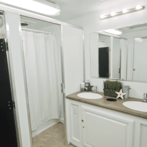 A modern bathroom with a large mirror above a double sink vanity, white cabinets, a white shower curtain, and decorative items under bright lights
