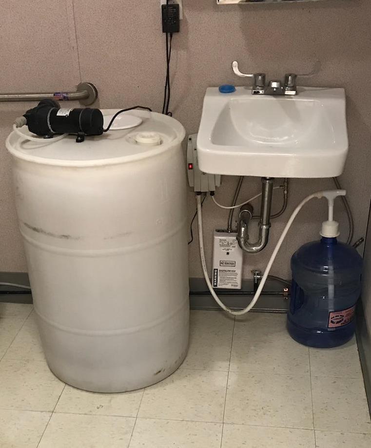 An ambient pump system sink consisting of a large white plastic barrel connected by hoses to a sink and a blue water jug