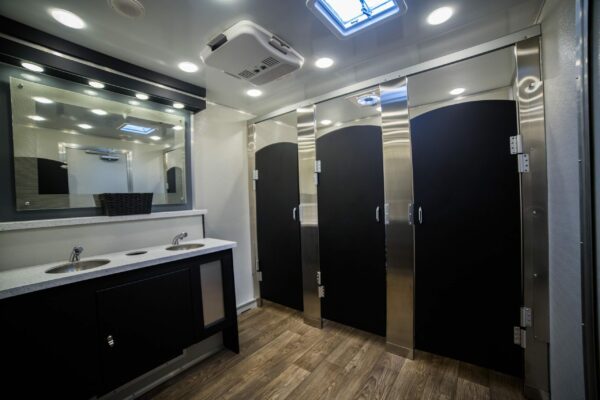 Interior of a modern mobile bathroom, featuring two closed stall doors, dual sinks with mirrors, wooden flooring, and overhead lighting