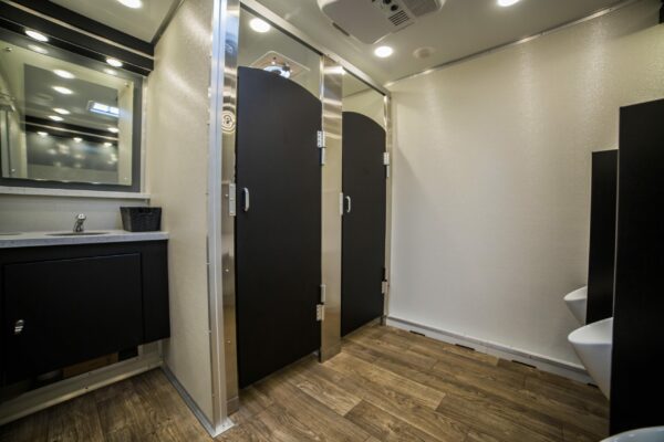 Interior of a modern mobile bathroom trailer featuring two closed stall doors, a sink with a black countertop, mirrors, and wood-like flooring