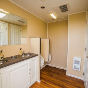 A clean trailer restroom with two sinks, mirrors, a partitioned area with urinals, and wooden flooring