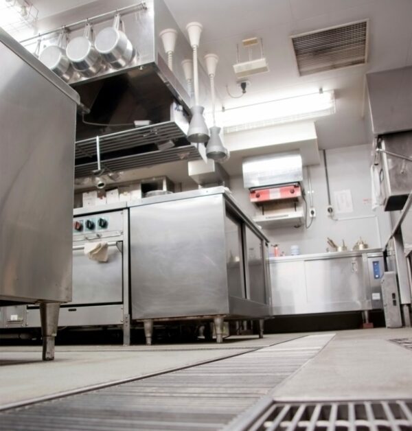 Professional restaurant kitchen interior showing a grease trap, stainless steel appliances, exhaust hoods, and floor grating, viewed from a low angle