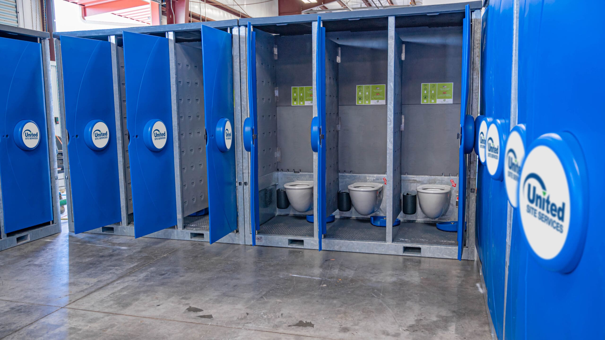 A row of open blue portable toilets, part of the USS Hydroflow System, in a large industrial space, each featuring a white toilet and hand sanitizer