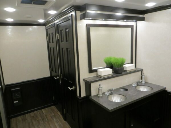 Interior of a restroom trailer featuring dual sinks with silver faucets, a large mirror, black cabinets, and a decorative green plant