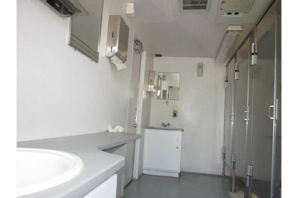 Interior of a clean portable restroom trailer featuring two sinks with mirrors above, hand dryers, and three urinals