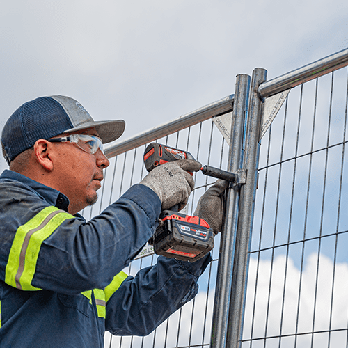 A worker in a high-visibility jacket and safety glasses is using a cordless drill to install a metal grid fence against a cloudy sky background