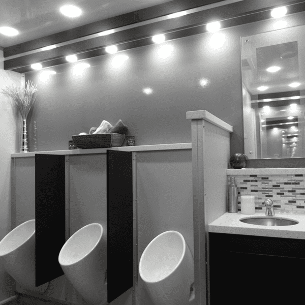 A modern mobile restroom interior featuring black stall doors, a stainless steel sink with a dark red towel, and a vase with red flowers on the counter