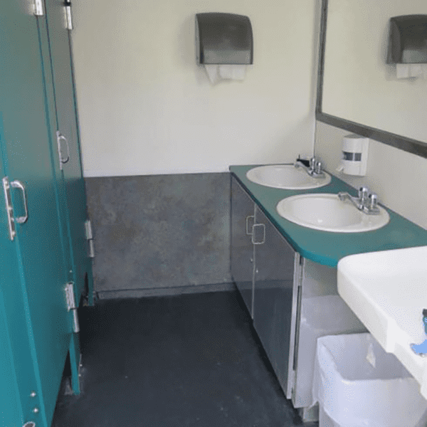 Trailer restroom interior with teal stall doors, two sinks with faucets, a toilet, and paper towel dispensers on wall tiles and gray flooring