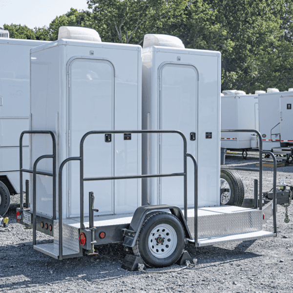 Two mobile solar-powered restroom trailers parked on a gravel lot, equipped with steps and railings, and each trailer connected to a small towing wheel assembly
