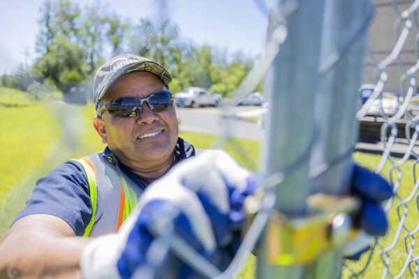 A smiling worker wearing a cap, sunglasses, and a reflective vest attaching a fence panel