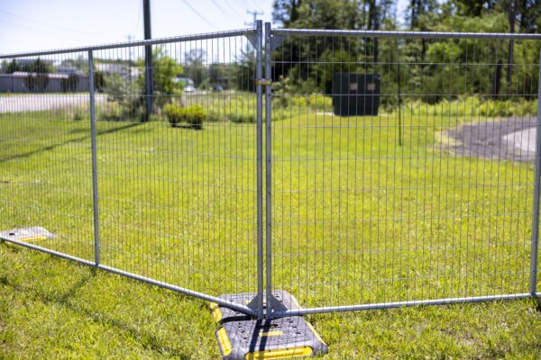 A temporary metal fence panel with a mesh grid pattern, set up on grass in a sunny outdoor area, stabilizing feet visible on the bottom with green trees and a paved path in the background