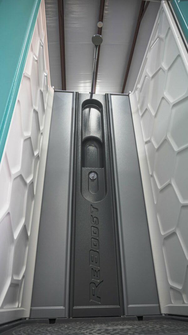 An interior view of a mobile shower unit, focusing on the textured gray door and latch with a hexagonal pattern on the walls, under artificial lighting