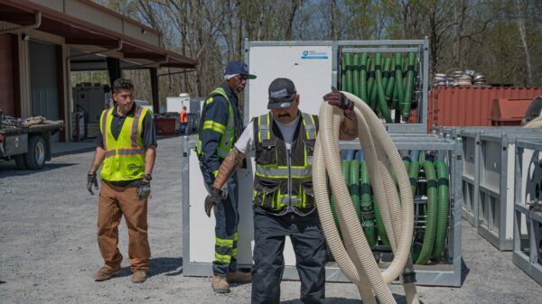 Three workers in high visibility vests at a worksite, handling and organizing large hoses stored on a metal rack outdoors