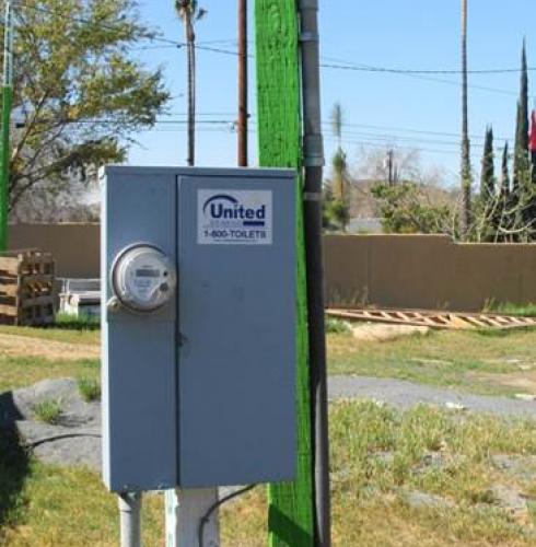 A gray temporary power box labeled "United Site Services" attached to a green wooden pole, set against a background of clear sky, grass, and shrubbery
